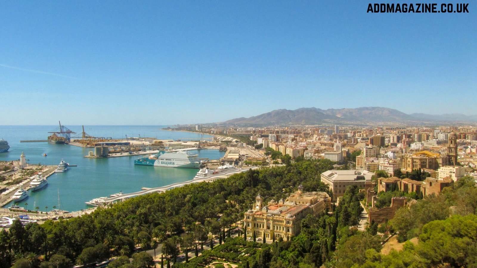 Elevated view of Malaga Port with a Balearia ferry, city landmarks, and mountains under a clear blue sky.